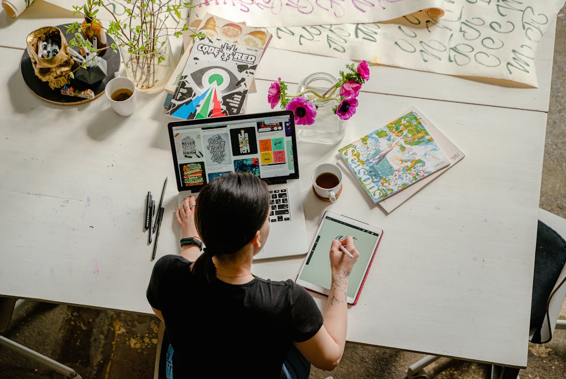 Personne créant du contenu numérique: une femme assise à une table blanche avec un laptop, une tablette graphique et un crayon, entourée de plantes, magazines et tasses, travaillant sur des designs.