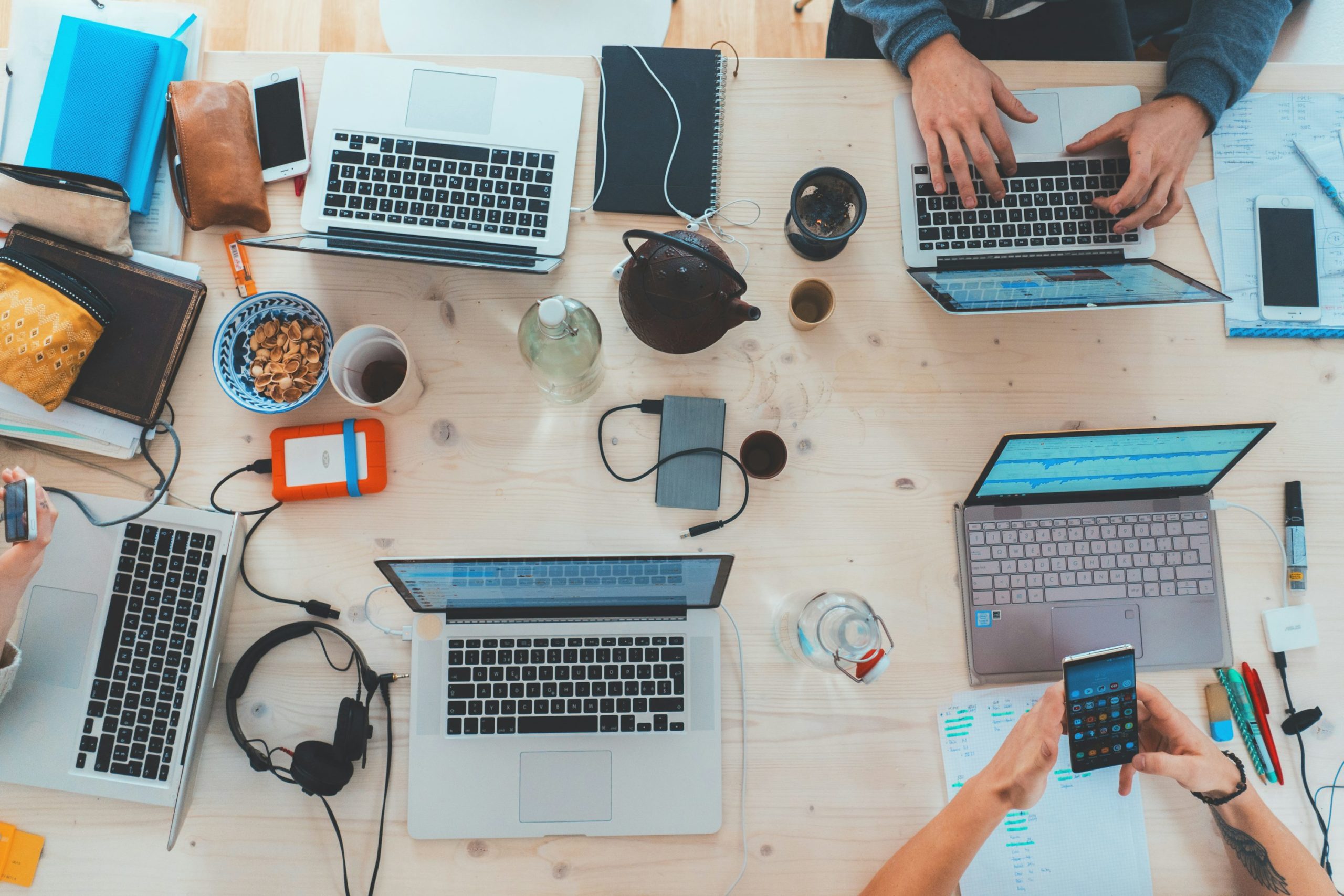 Équipe de travail en coworking avec plusieurs ordinateurs portables, smartphones et accessoires sur une table, collations et tasses.